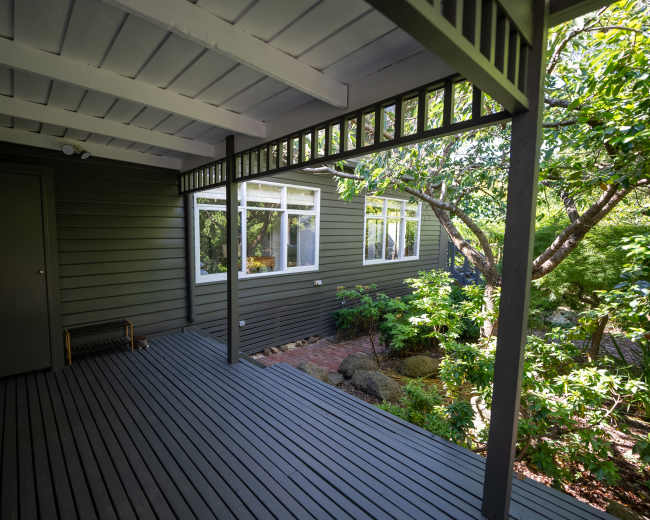 A shaded porch with wooden flooring leads to a house featuring green siding and large windows surrounded by lush foliage.