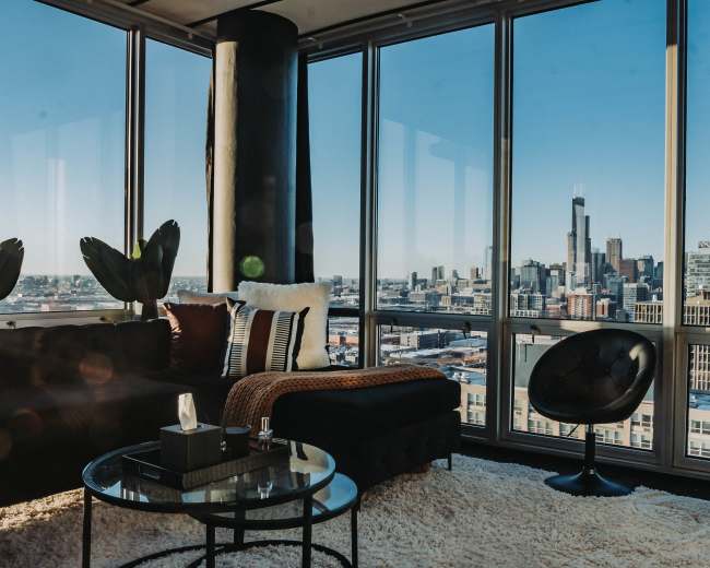 A modern living room features a large window showcasing a city skyline, with a black sofa, a round chair, and a shaggy rug.
