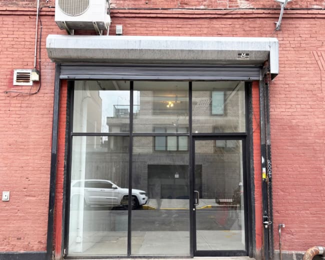 The image shows a vacant storefront with large glass panels and a metal awning, located on a brick wall.