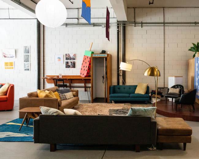 The image shows a spacious lounge area with various seating arrangements, including a green couch, a brown sofa, and a wooden table, surrounded by bookshelves and decorative items.