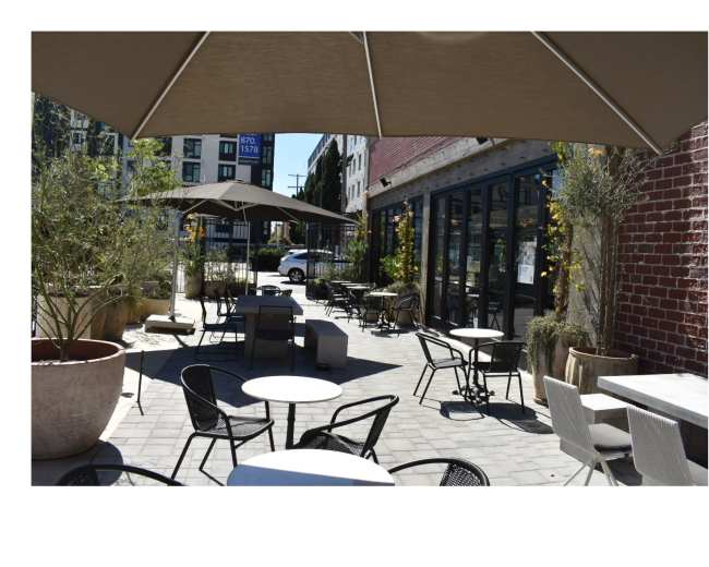 A patio area is set up with metal and wooden furniture, shaded by large umbrellas, and surrounded by planters and a brick wall.