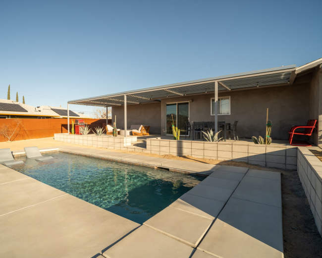 A modern house with a pool is situated in a desert landscape, featuring a shaded patio area and minimal landscaping.