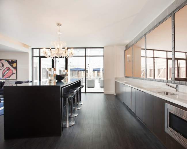 The image shows a modern kitchen with a bar counter, high stools, and large windows allowing natural light to enter the space.