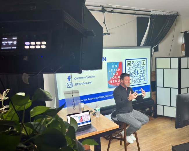 A speaker presents in front of a large screen in a well-lit room equipped with cameras and studio lighting.