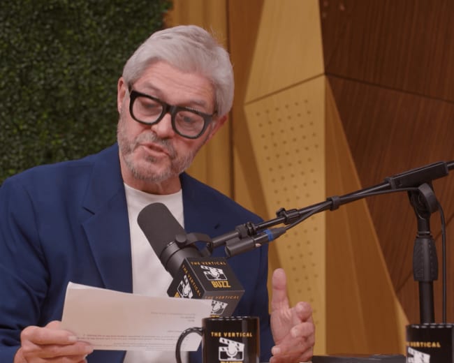 A man with gray hair and glasses reads from a paper while seated at a table with microphones and coffee cups.