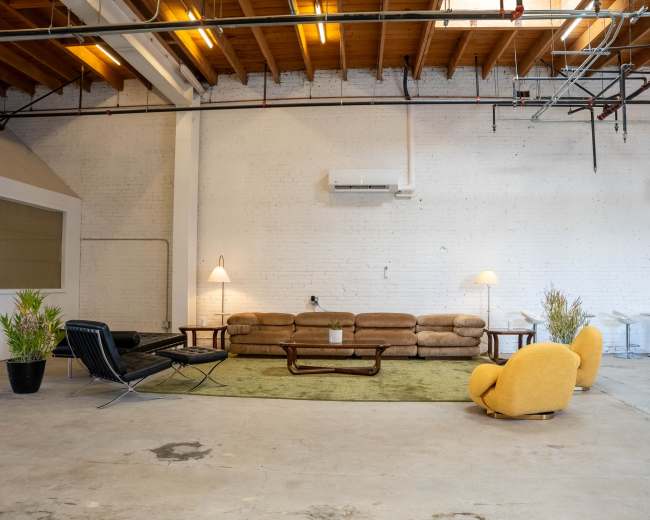 The image shows a spacious industrial-style room featuring a brown couch, a black lounge chair, yellow armchairs, and a green area rug, with exposed wooden beams and air conditioning units on the brick walls.