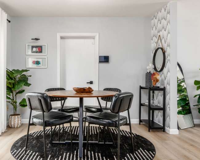A modern dining area features a round table surrounded by four black chairs, with a plant and decorative items positioned against light gray walls.