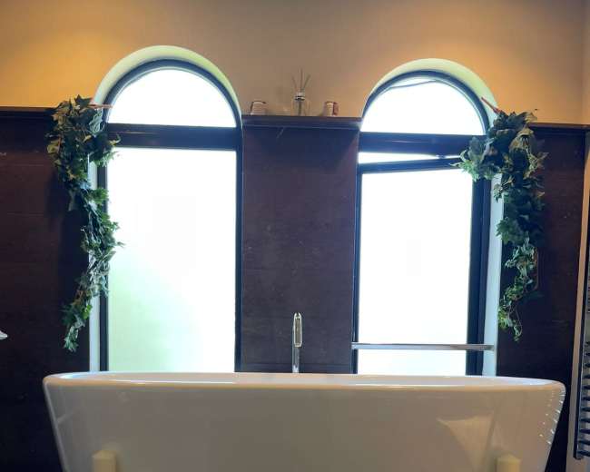A modern freestanding bathtub is positioned between two arched windows adorned with greenery, flanked by candles on ornate holders.