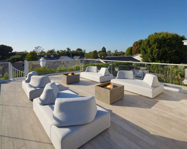 A modern rooftop terrace features several white lounge chairs and fire pits surrounded by greenery and clear skies.