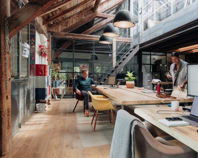 A bright, industrial-style office features wooden beams, large windows, and a long table with a person working at a computer while another prepares coffee.