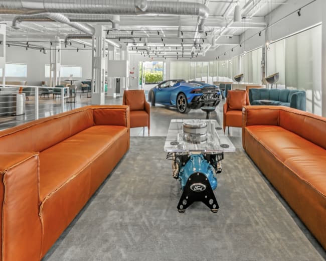 A modern lounge area with two leather sofas and a glass coffee table, featuring a blue sports car visible in the background.