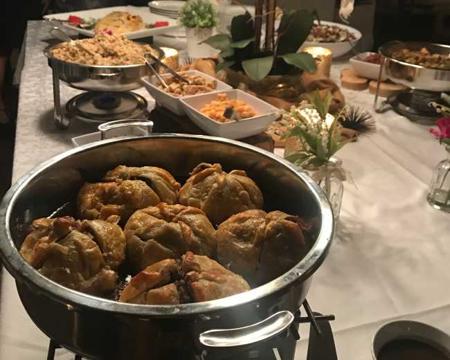 A buffet table is set up with a variety of dishes, including a large pot of baked dumplings and garnishes, surrounded by guests and decorative flowers.