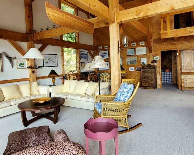 The interior of a rustic cabin features wooden beams, a spacious living area with a sectional sofa, a rocking chair, and a small pink table, all illuminated by natural light.