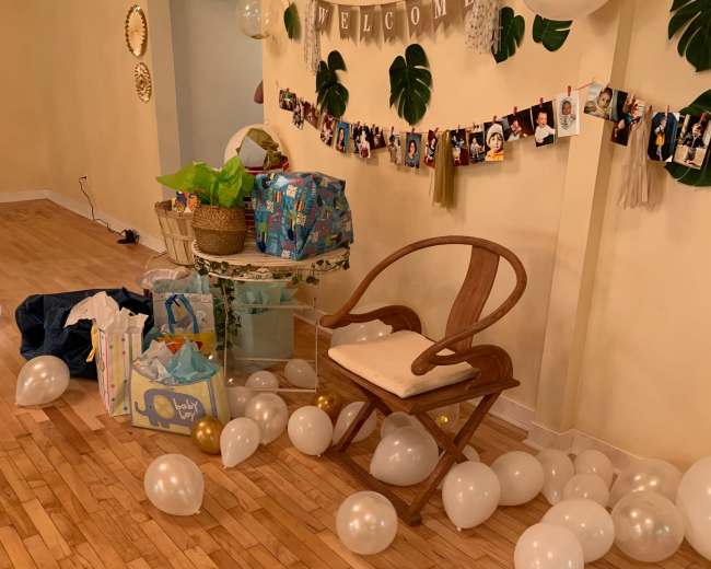 A room decorated for a celebration features a welcome banner, photos hanging on a string, a chair, and scattered white balloons on the wooden floor.