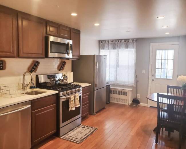 The image shows a kitchen with wooden cabinets, stainless steel appliances, a small dining table, and a door leading to the outside.