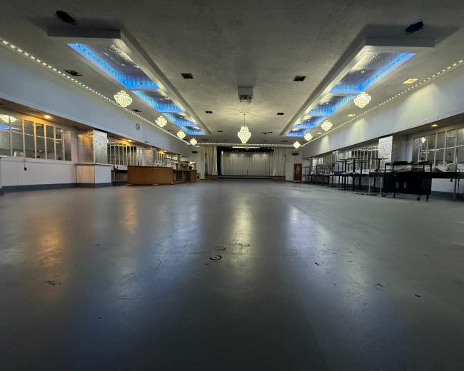 The image shows a large, empty hall with a polished floor, overhead chandeliers, and blue lighting accents along the ceiling.