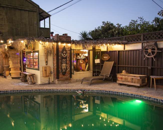 A backyard scene featuring a pool surrounded by a tiki-style bar, string lights, and a seating area within a tropical setting.