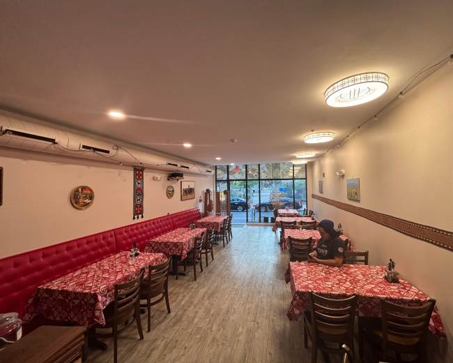 The image shows the interior of a restaurant with red tablecloths, wooden chairs, and large windows providing natural light.