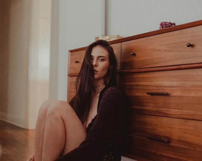 A person sits on the floor next to a wooden dresser in a room with warm lighting.