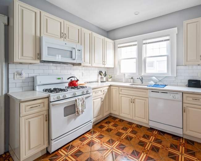 The kitchen features cream-colored cabinets, a gas stove, a microwave, and a dishwasher, with a tiled floor pattern and ample natural light from the windows.