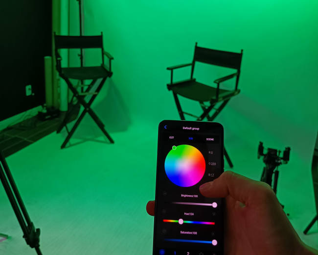 A person holding a smartphone displaying a color adjustment tool in front of two empty director's chairs in a green-lit studio setting.