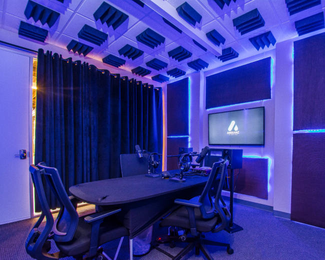 A soundproof recording studio features a circular table with microphones and chairs, illuminated by blue LED lights, and lined with acoustic panels.