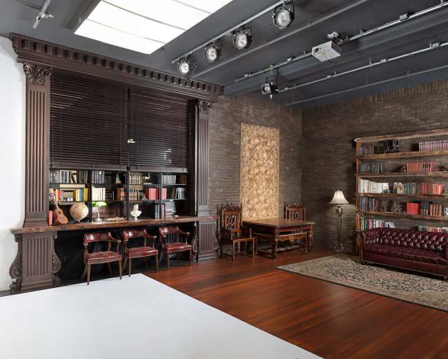 The image shows a modern studio space featuring a wooden desk and chairs, a bookshelf filled with books, a leather couch, and a plant, all arranged against a brick wall.