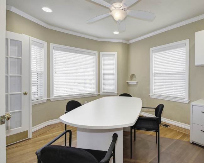 A brightly lit room features a large white table surrounded by black chairs, with multiple windows allowing natural light to enter.