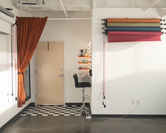 The image shows a contemporary studio space with a barstool near a checkered floor and colorful backdrop rolls mounted on the wall.