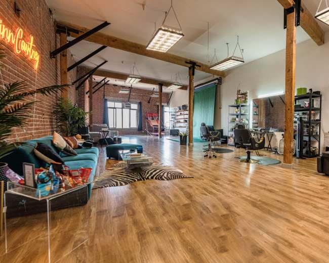 A spacious, modern studio with wooden flooring, large windows, and a mix of seating areas, including a teal sofa and several salon-style chairs.