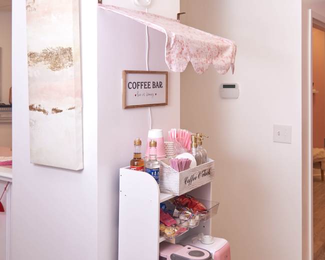 The image shows a coffee bar setup in a small nook, featuring a pink coffee machine, colorful drink supplies, and decorative signage.