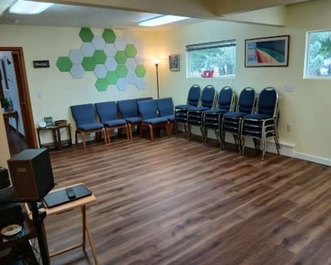 The image shows a room with wooden flooring, a row of blue chairs against the wall, and a stack of chairs in the corner.