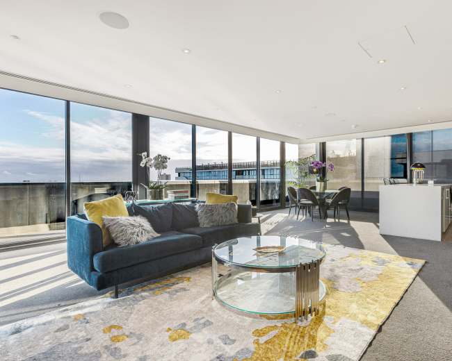 The image shows a modern living room with large floor-to-ceiling windows, a blue sofa, a circular glass coffee table, and a dining area with dark chairs and an open kitchen in the background.