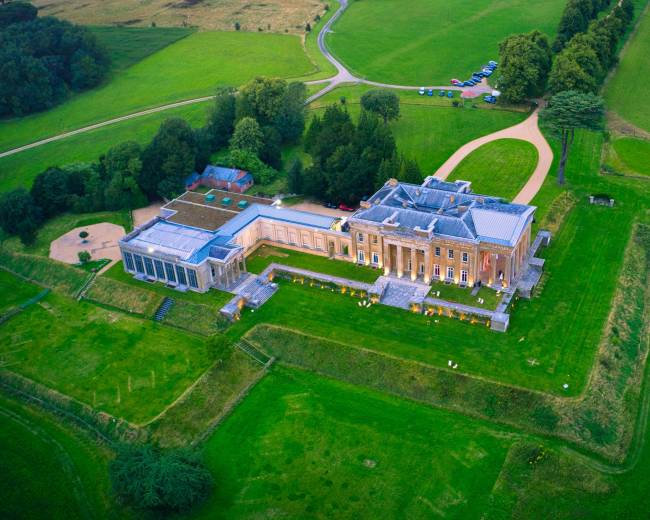 The image shows a large mansion situated on a sprawling green landscape with a winding road leading to it, surrounded by trees and open fields.