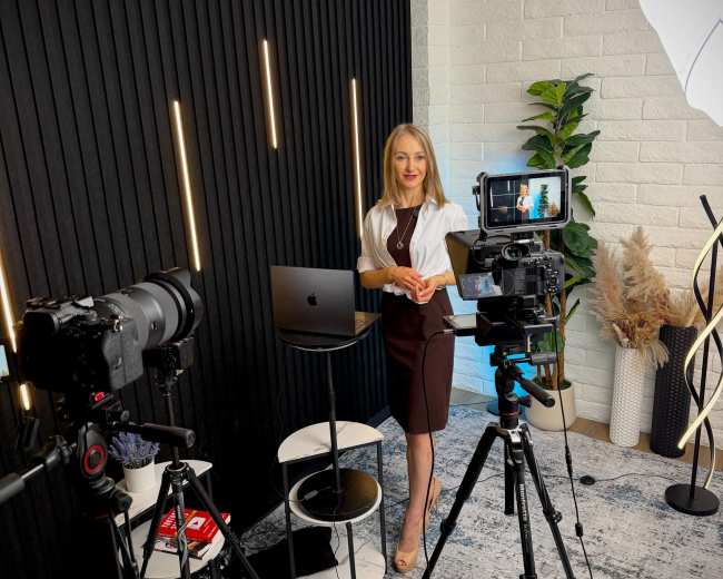 A woman stands in a studio space with multiple cameras set up, a computer on a table, and decorative plants in the background.