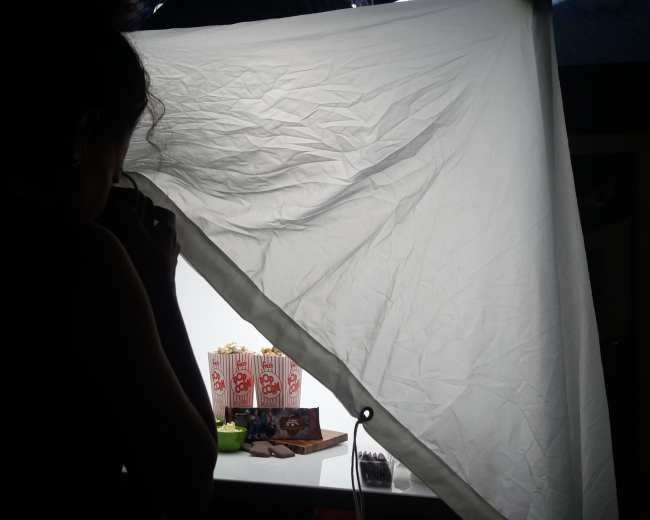 A person is taking a photograph of objects arranged on a table, with a large white backdrop and a softbox light source set up in the scene.