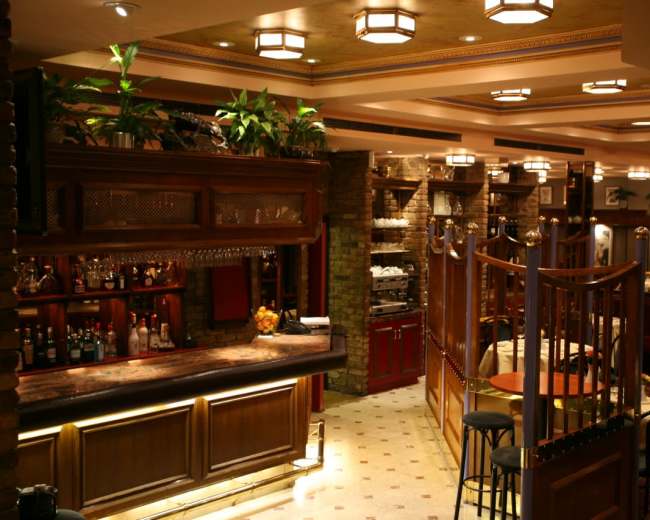 An elegant restaurant interior featuring a wooden bar, various plants, and separate dining areas with round tables and chairs.