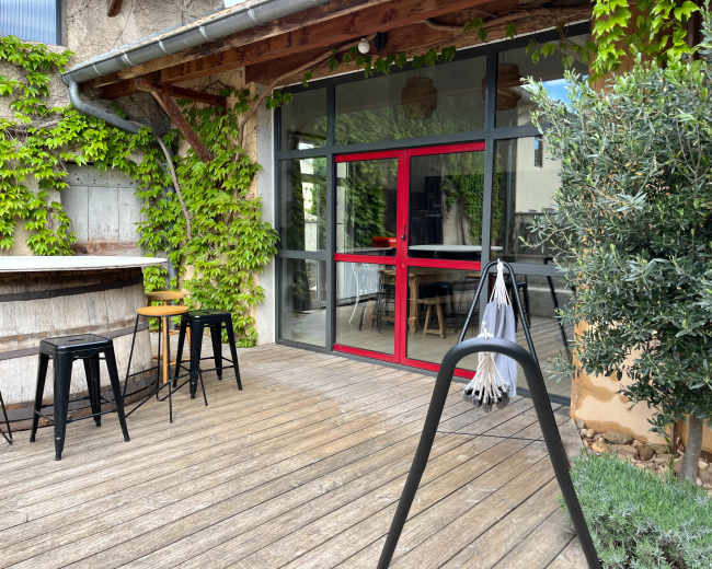 The image shows a wooden deck with black stools, a swing, and a glass door framed in red, surrounded by greenery.