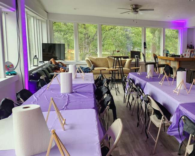 The image shows a spacious indoor area set up with several rows of purple-covered tables and easels, equipped for an art activity, alongside a bar and a seating area.