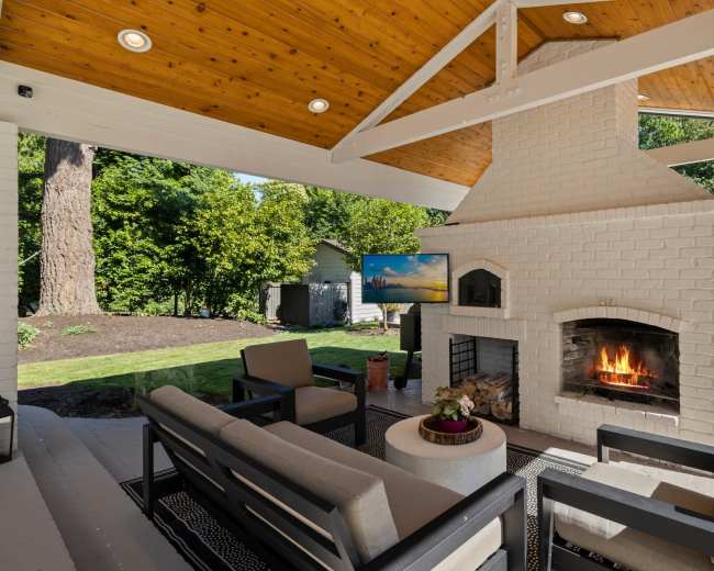 A covered outdoor seating area features a brick fireplace, comfortable chairs, and a television against a backdrop of greenery.