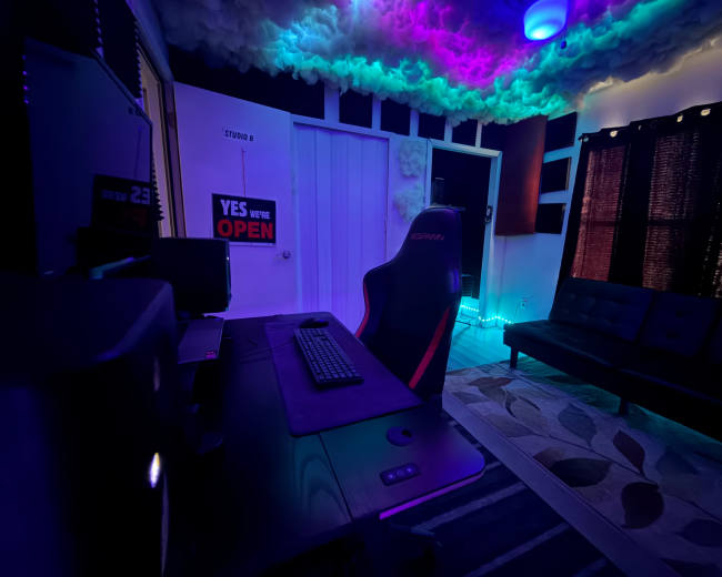 A gaming setup features a sleek black desk with a keyboard and monitor, surrounded by colorful lighting and a cozy seating area with a black couch.