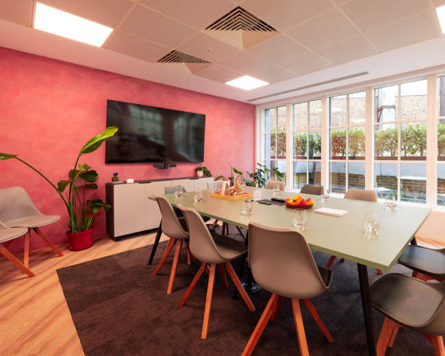 A modern conference room features a long table surrounded by chairs, a large TV on the wall, and windows overlooking a green outdoor space.