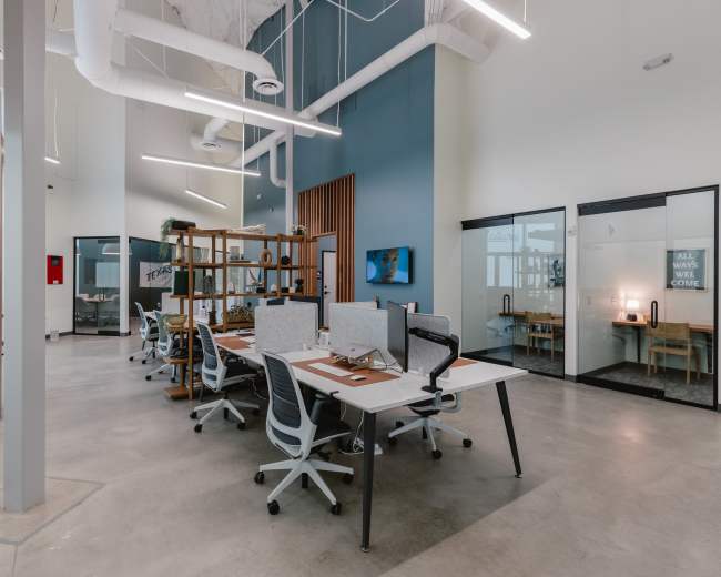 The image depicts a spacious office environment with modern desks and chairs arranged in a layout that encourages collaboration, featuring glass-walled meeting rooms and a neutral color scheme.