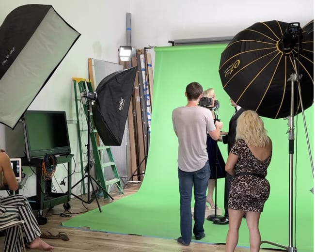 A photography studio features a green backdrop, multiple lighting equipment, and people preparing for a photoshoot.