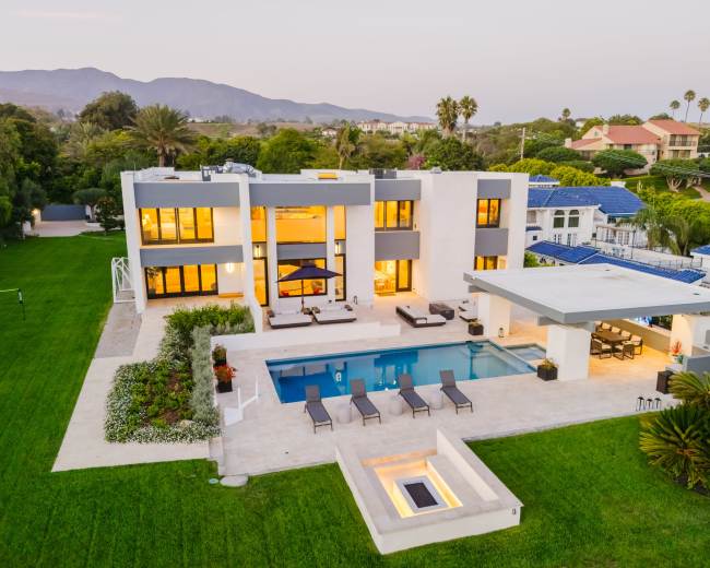 A modern two-story house with large windows, a swimming pool, and a landscaped yard is located in a suburban area with mountains in the background.