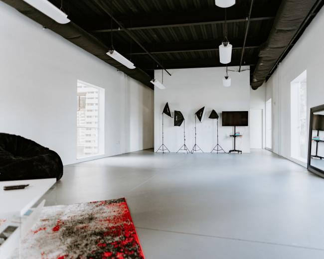 An empty, modern studio space features white walls, large windows, three light stands, and a television on a stand.