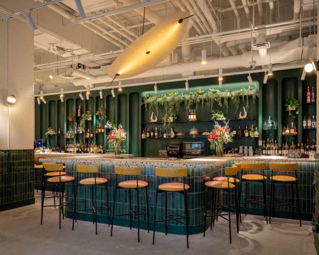 The image shows a modern bar with a green and gold color scheme, featuring a curved counter, high stools, and shelves stocked with various bottles, adorned with plants and decorative elements.