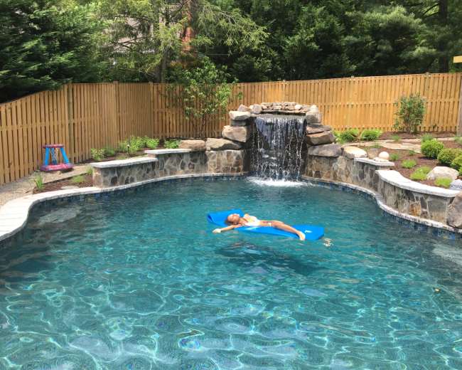 A girl floats on a blue pool float in a backyard swimming pool with a stone waterfall and a wooden fence in the background.