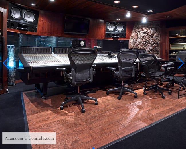 A control room with various audio equipment and monitors, featuring a large mixing console at the center and multiple chairs arranged for operators.