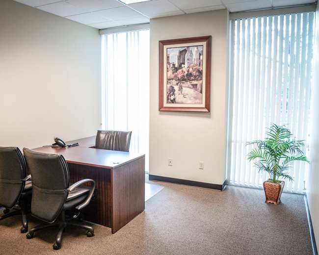 The image shows a simple office space with a wooden desk, two black chairs, a telephone, and a framed painting on the wall, along with a plant in a pot.